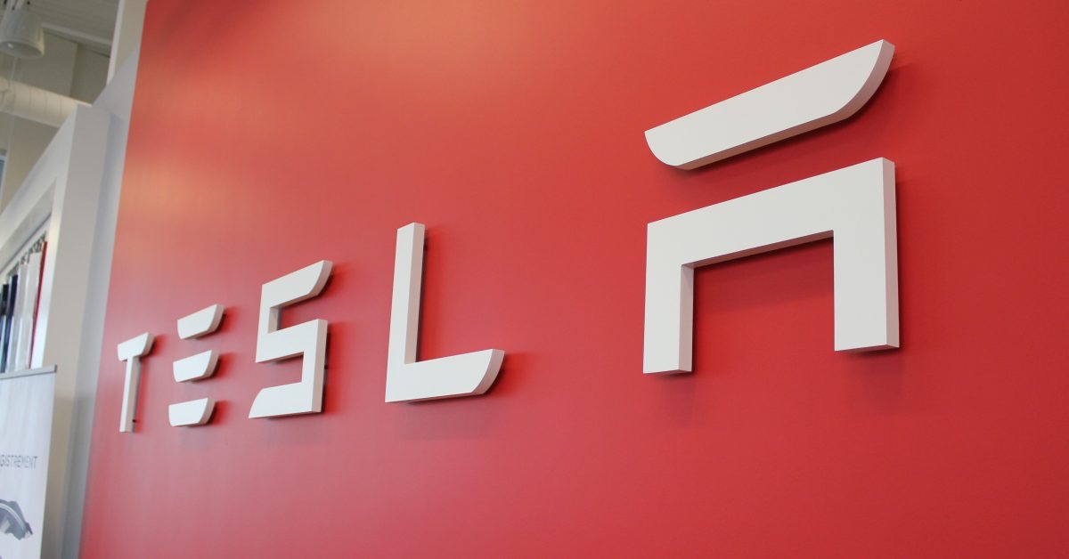 White Tesla lettering and logo mounted on a red showroom wall, used as the ThriveInMarkets evening-review inline image for Friday October 2 2026, when Tesla closed 374.16 dollars up 5.7 percent after third-quarter deliveries of 486,532 vehicles beat a 461,974 consensus