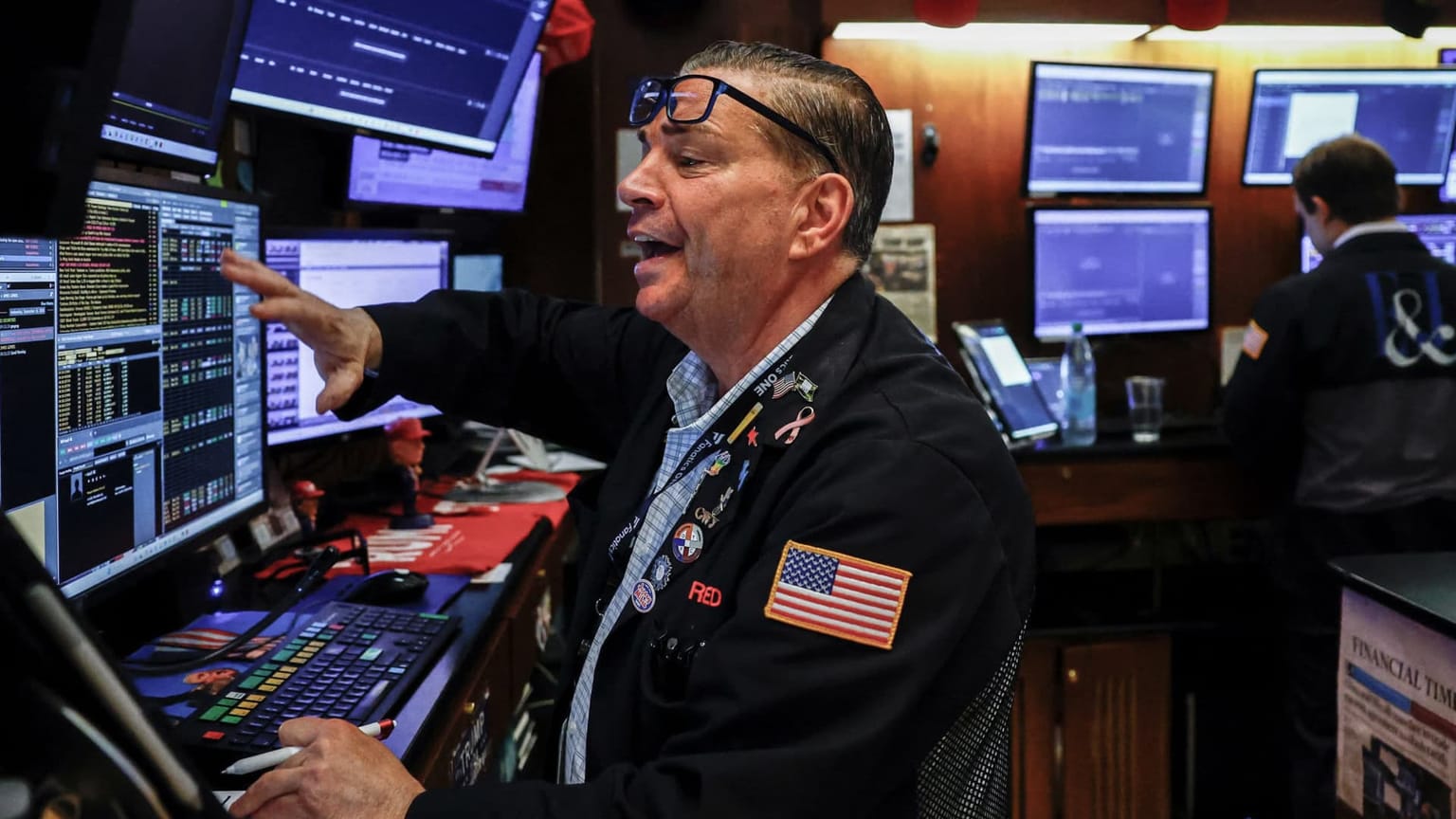 A New York Stock Exchange floor trader in a black jacket points at quote screens, used as the ThriveInMarkets evening-review inline image for Monday October 5 2026, when PTC jumped about 34 percent after Schneider Electric agreed a 22.6 billion dollar cash bid and the Nasdaq Composite closed 27,477.31 at a record