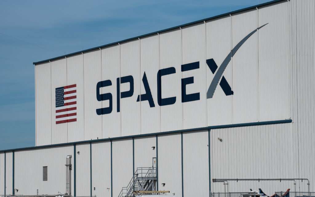 The exterior of a large white SpaceX hangar building displaying the SpaceX company logo and a United States flag under a clear blue sky, illustrating the ThriveInMarkets coverage of SpaceX joining the Nasdaq-100 index before the open on Tuesday July 7 2026, a fast-tracked inclusion expected to trigger roughly 4.3 billion dollars of forced passive buying from Nasdaq-100 trackers such as the Invesco QQQ and QQQM against a small publicly traded float, as the S&P 500 sits at a record 7,537.43 and megacap technology extends its rebound from late June's Meta Compute shock.