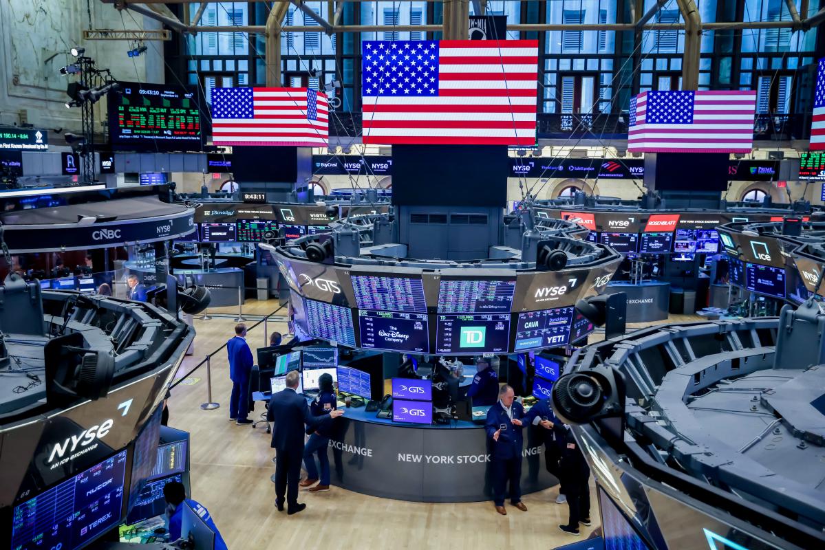 The New York Stock Exchange trading floor with US flags over the GTS booths and quote screens, used as the ThriveInMarkets week-ahead hero for October 19 to 23 2026, when Tesla and Meta report after the close on Wednesday and Friday's S&P Global flash PMIs are the last US activity print before the October 27-28 FOMC