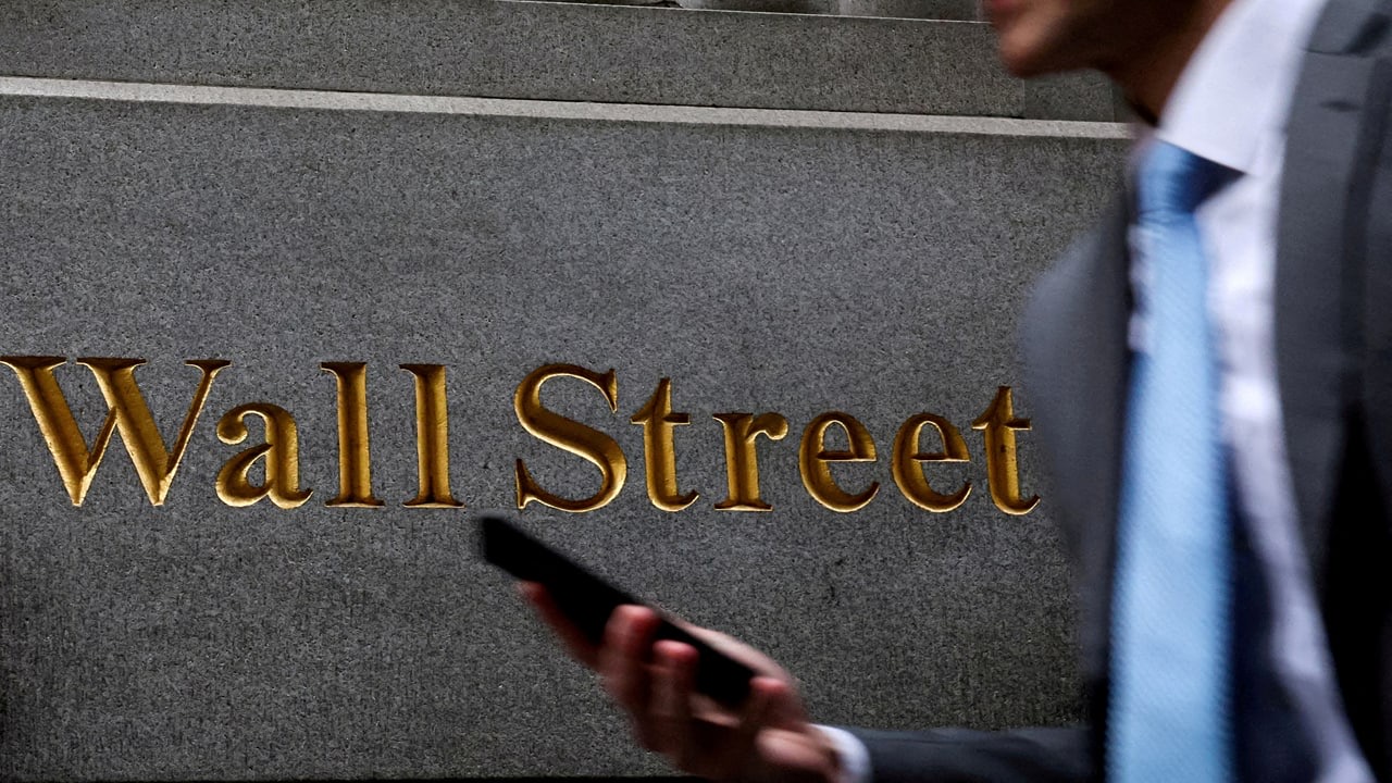 A pedestrian in a grey suit and light-blue tie walks past the gold Wall Street inscription on a granite facade, used as the ThriveInMarkets weekly markets roundup hero for August 23 to 29 2026, when Chair Kevin Warsh's first Jackson Hole keynote lifted September hike odds toward 58 percent, Nvidia guided 70 percent fiscal 2028 revenue growth, Salesforce jumped about 23 percent, Solana passed a double-disinflation vote, and the S&P 500 closed the week at 7,711.76